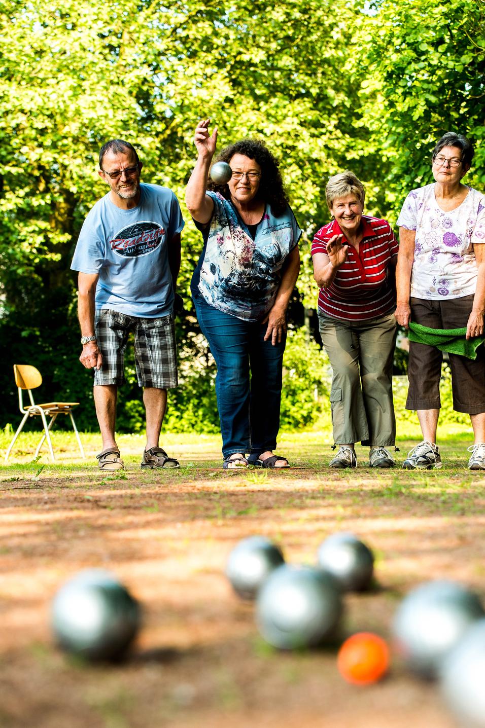 Vier Personen spielen Boule im Freien, während eine Frau gerade eine silberne Kugel wirft.