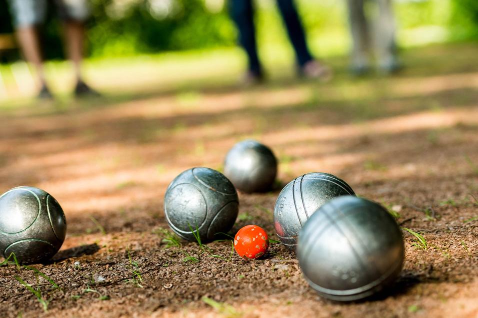 Gleise mit silbernen Boulekugeln und einer roten Kugel auf einem erdigen Boden in einem Park.
